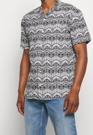 Homme portant une chemise à manches courtes noire et blanche à motifs géométriques et un jean bleu clair, debout sur un fond blanc uni.