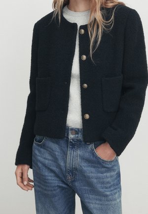 Woman wearing black textured button-up jacket over light gray top and blue denim jeans with one hand in pocket against neutral background.