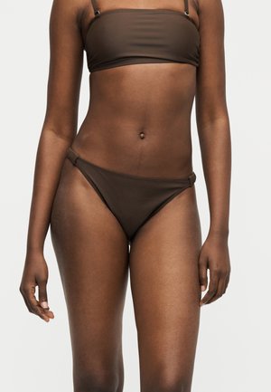 Žena oblečená v tmavohnedom dvojdielnom bikinách s bandeau vrchným dielom a nohavičkami s nízkym pásom na jednoduchom pozadí.