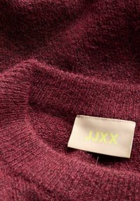 Bordo megzto džemperio medžiaga su minkštu, tekstūruotu paviršiumi. Apima šviesiai smėlio spalvos etiketę su "JJXX" geltonai.