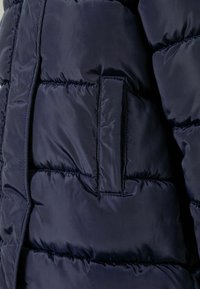 Tmavomodrá prešívaná bunda s hladkým, lesklým povrchom a prešívanými časťami, s detailom bočnej vrecka.