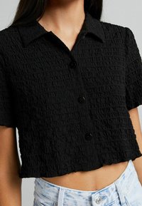 Camicetta corta a maniche corte nera con texture, colletto e bottoni, indossata con jeans a vita alta in denim chiaro effetto acido.