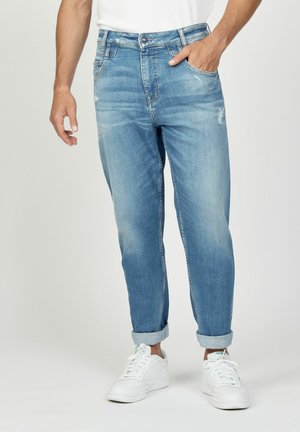Mann mit hellblauen hochgekrempelten Jeans und weißen Sneakern, steht mit einer Hand in der Tasche vor einem einfarbigen Hintergrund.