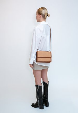 Donna in piedi, indossa una camicia bianca oversize, minigonna beige, stivali neri al ginocchio e una borsa a tracolla color cuoio con trama intrecciata.