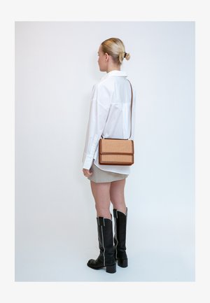 Femme debout, portant une chemise blanche oversize, une mini-jupe beige, des bottes noires montantes et un sac bandoulière marron avec une texture tissée.