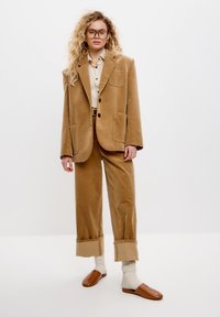 Blazer in velluto a coste marrone con grandi tasche, camicia beige e pantaloni a gamba larga abbinati. Scarpe marroni slip-on e calze crema completano il look.