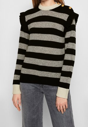 Vrouw draagt een zwart-grijs gestreepte trui met witte manchetten, schouderruche en gouden knopen, gecombineerd met grijze jeans.