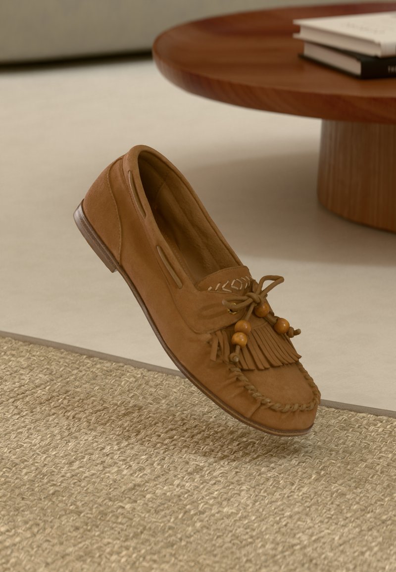 Mocassin en daim marron avec des franges, des perles décoratives et un bord tressé. Caractérisé par un bout rond et un faible talon en bois.
