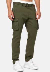 Saarenvihreät työhousut sivutaskuilla, säädettävällä vyöllä, kapealla leikkauksella ja vahvistetuilla saumoilla, yhdistettynä valkoisiin lenkkareihin.