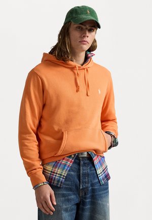 Haut à capuche orange avec une poche avant, associé à une chemise à carreaux bleue et à boutons. Un jean bleu foncé complète la tenue, avec une casquette en velours côtelé verte.
