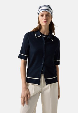 Woman wearing a navy blue short-sleeve buttoned cardigan with white trim, white pants, and a white and navy striped headscarf.