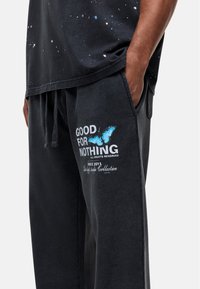 Zwarte trainingsbroek met een vlinderafbeelding en de tekst "GOED VOOR NIETS." Zacht materiaal met een elastische tailleband en zijzakken.