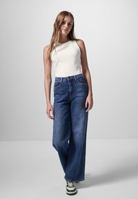 Witte mouwloze top gecombineerd met hoge taille, wijde donkerblauwe jeans. Model draagt groene en witte sneakers. Effen achtergrond.
