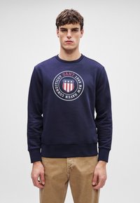 Granatowy bawełniany sweatshirt z okrągłym dekoltem, ściągaczami na rękawach i elastycznym wykończeniem. Z przodu znajduje się wyraźny okrągły motyw z logo GANT.