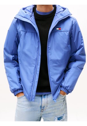 Mann trägt hellblaue Kapuzenjacke über schwarzem Pullover und hellblaue zerrissene Jeans, Hände in den Taschen, Vorderansicht, über dem Mund beschnitten.