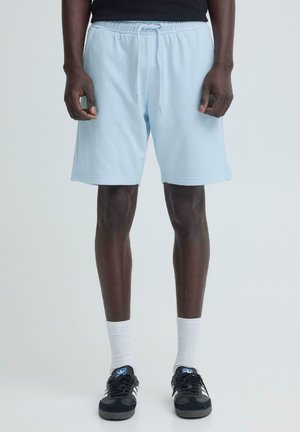 Mees, kes kannab helehalli paelaga lühikesi pükse, valgeid tavalisi sokke ja musti Adidas tossusid, seisab tavalise heleda tausta ees.