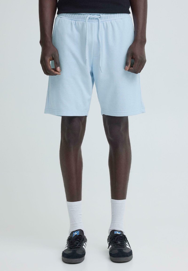 Άνδρας που φοράει ανοιχτό μπλε σορτς με κορδόνια, λευκές κάλτσες μέχρι τον αστράγαλο και μαύρα αθλητικά παπούτσια Adidas, στέκεται μπροστά σε ένα απλό ανοιχτό φόντο.