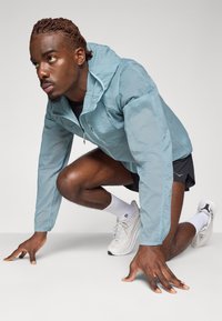 Lichtblauwe, lichtgewicht jas met capuchon en rits aan de voorkant, gedragen over een zwart shirt en shorts. Witte sportschoenen en sokken zijn zichtbaar.