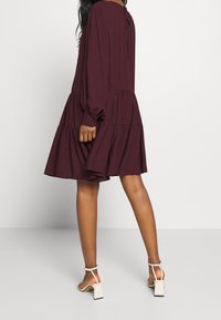 Burgundská šaty ve tvaru věže s dlouhými nafouknutými rukávy a detaily ve tvaru klíčové dírky vzadu, s volným siluetou. V kombinaci s bílými sandály na blokovém podpatku.