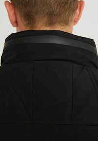 Ansicht von hinten einer Person mit kurzen Haaren, die eine schwarze Jacke mit hohem Kragen trägt, der mit einem Gummistreifen und einem dezenten gesteppten Design versehen ist.