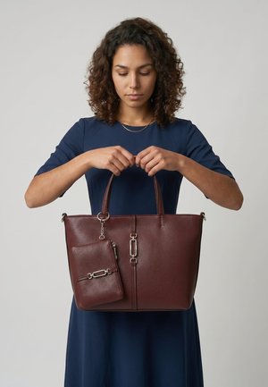 Frau mit lockigem Haar, die ein blaues Kleid trägt und eine große braune Lederhandtasche mit einer passenden angebrachten Tasche hält.