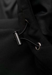 Tessuto nero con chiusura a coulisse, dotato di un toggles lucido nero e occhiello argentato, che mette in evidenza una superficie liscia e testurizzata.