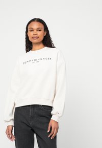Lys beige genser med ribbestrikkede mansjetter og nederkant, med svart logo-tekst "TOMMY HILFIGER EST. 1985" over brystet. Kombinert med mørke jeans.