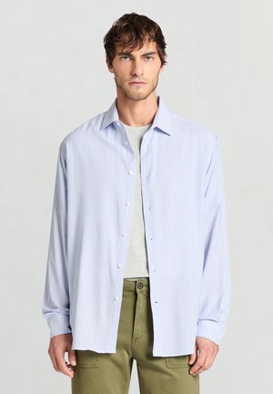 Hellblau gestreiftes Langarmhemd mit Knopfleiste, weichem Stoff, lockerem Schnitt, übereinander getragen über einem grauen T-Shirt, kombiniert mit olivgrünen Hosen.