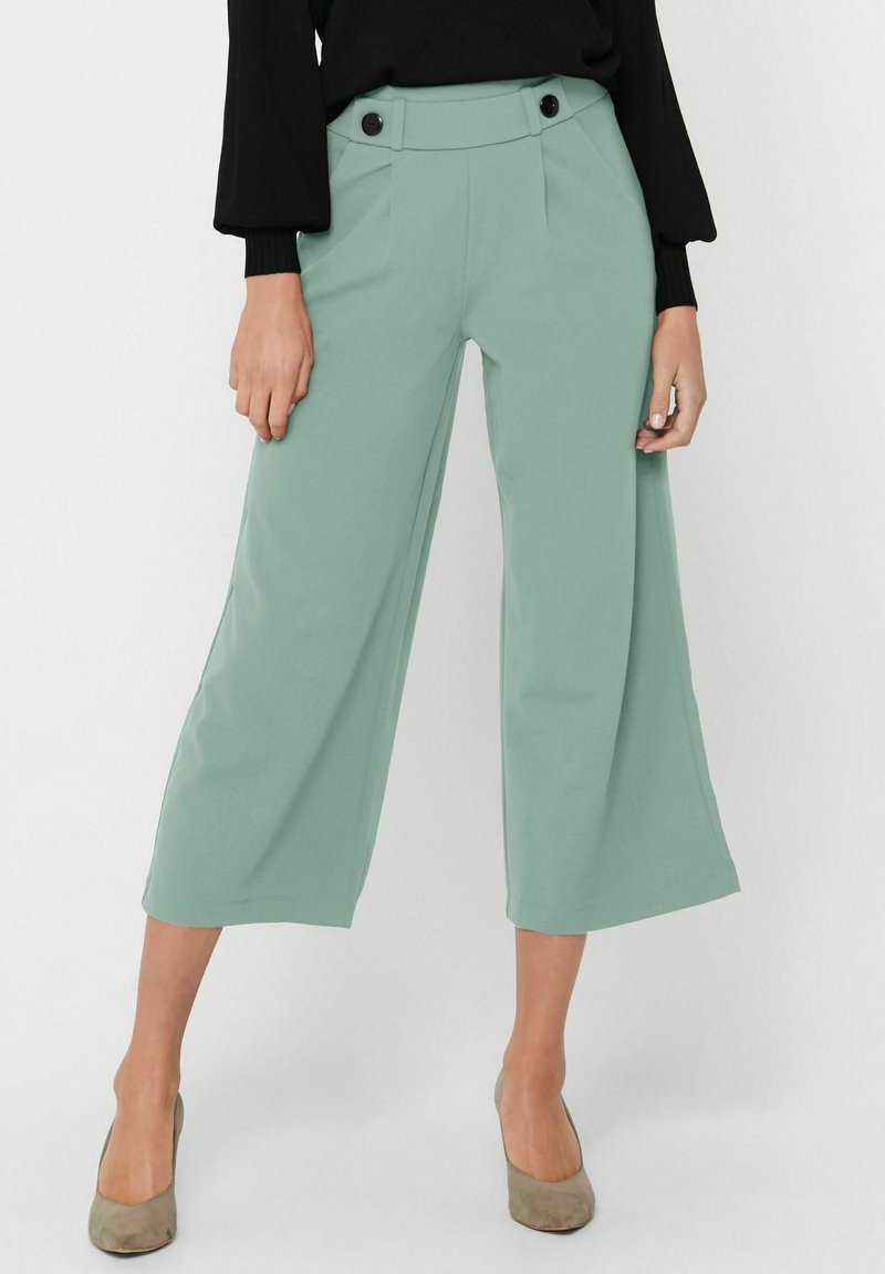 Pantaloni ampi di color verde menta con pieghe e due bottoni neri in vita. Tessuto liscio, lunghezza cropped, abbinati a tacchi neutri.
