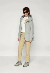 Heled hall veekindel jakk kapuutsiga, beežid püksid ja hallid matkajalatsid. Omab lihtsat disaini, esipockets ja tõmblukku.