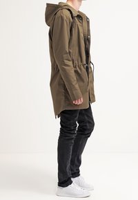 Olivově zelená parka s kapucí a staženým pasem, zapínáním na knoflíky a zip a bočními kapsami, kombinovaná s černými džíny a bílými teniskami.