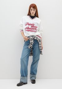 Sweatshirt bianco oversize con testo grafico rosso, abbinato a jeans blu a gamba larga. Una camicia scozzese annodata evidenzia la vita, e stivaletti neri con suola spessa completano il look.