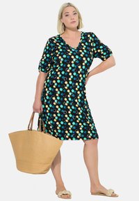 Schwarzes Kleid mit großen blauen, türkisfarbenen, gelben und weißen Punkten; V-Ausschnitt, kurze Ärmel; kombiniert mit einer gewebten beigen Tragetasche und Sandalen.