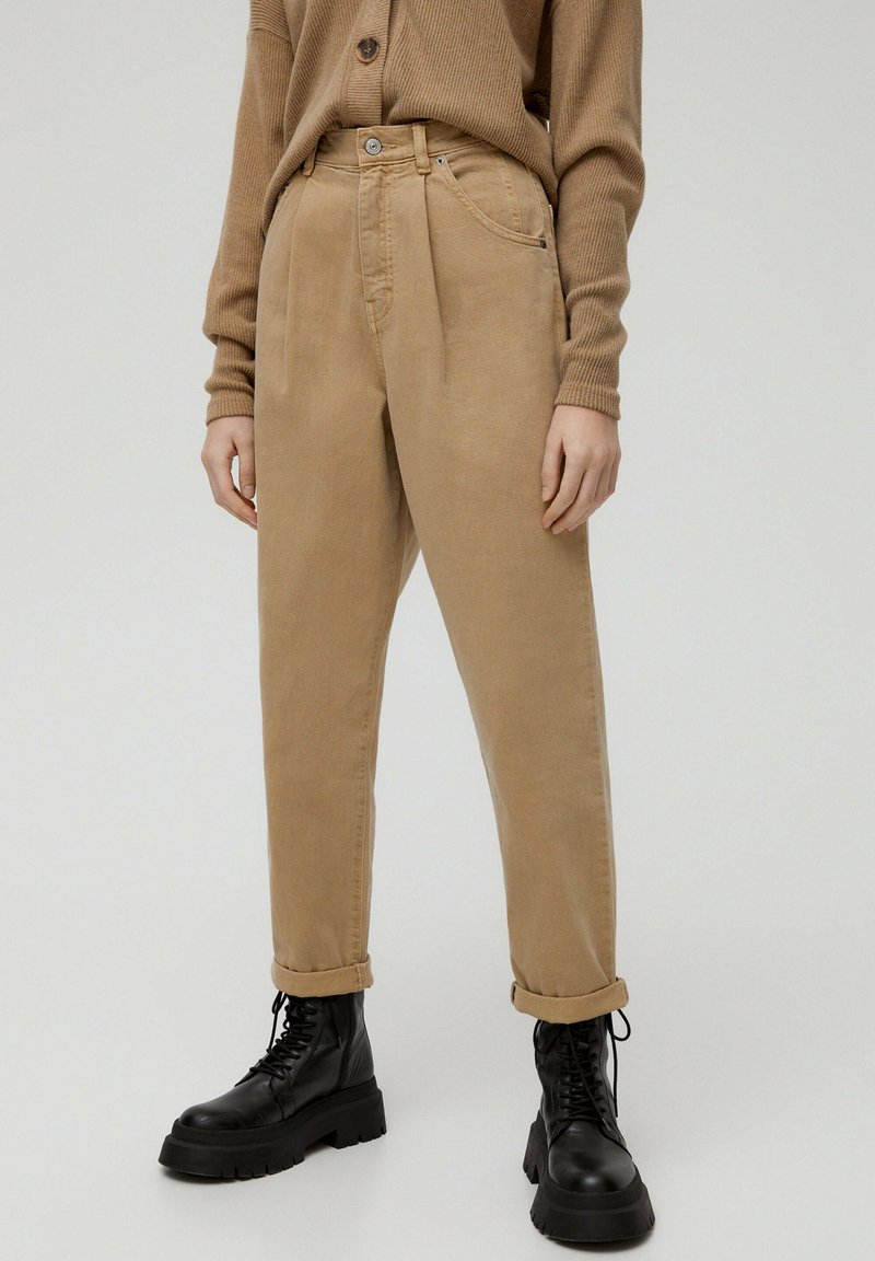 Personne portant un pantalon beige retroussé, un pull côtelé marron et des bottes noires épaisses à lacets, debout devant un fond clair uni.