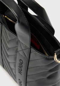 Bolso de mano de cuero negro acolchado con cremallera dorada, correa ancha para el hombro y la marca "HUGO" en la correa y el forro interior.