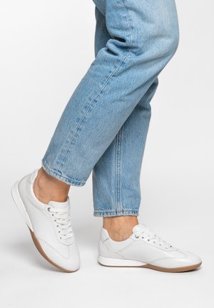 Personne portant un jean bleu clair et des baskets blanches à lacets avec des semelles marron, un pied légèrement levé, sur un fond uni.