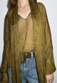 Gennemsigtig olivengrøn bluse med et struktureret slange mønster og et draperet design. Parret med højtaljede blå jeans og et brunt bælte.