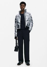 Giubbotto puffer argentato con design trapuntato, colletto alto e polsini elastici, abbinato a pantaloni scuri e borsa nera. Scarpe testurizzate completano il look.