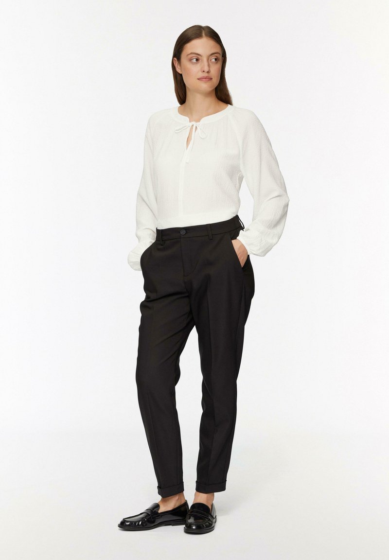 Blouse blanche texturée avec un décolleté en trou de serrure et un col à nouer, manches longues, associée à un pantalon noir ajusté et des mocassins noirs. Design simple et épuré.