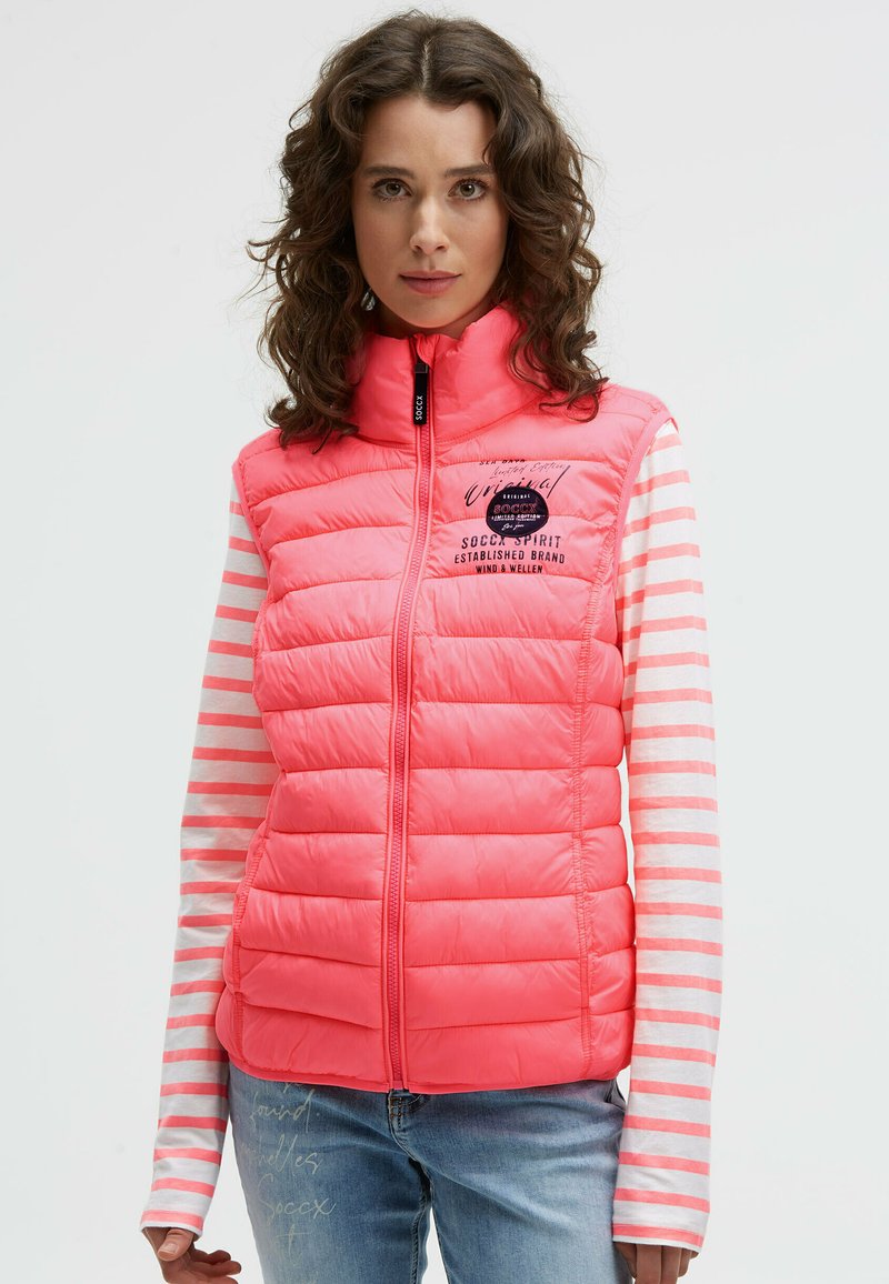 Pink puffer vest with a high collar and zip closure. Features quilted design and a logo on the chest. Worn over a striped long-sleeve top.