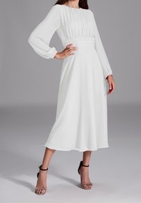 Robe blanche à manches longues avec taille cintrée, jupe fluide et texture douce. Accompagnée de talons noirs à bride de cheville et d'un fond neutre.