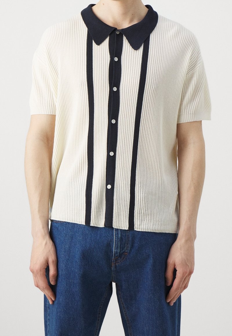 Homme portant un polo en maille crème à manches courtes avec des rayures verticales noires et un col, associé à un jean bleu, visible du cou aux hanches.