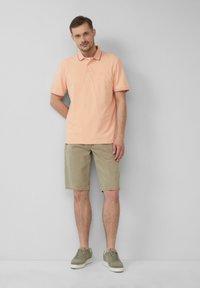 Jasnopomarańczowa koszulka polo z kołnierzykiem i kontrastowym wykończeniem, w połączeniu z beżowymi szortami i zielonymi sneakersami, stojąca na szarym tle.