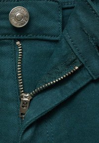 Tirkizni denim materijal s srebrnim dugmetom i metalnim zatvaračem. Krupni plan detalja zatvarača koji prikazuje zube i teksturu materijala.