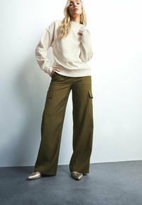 Beige sweatshirt med avslappnad passform, matchad med olivgröna cargobyxor med fickor. Modell som bär metalliska spetsiga ballerinas.