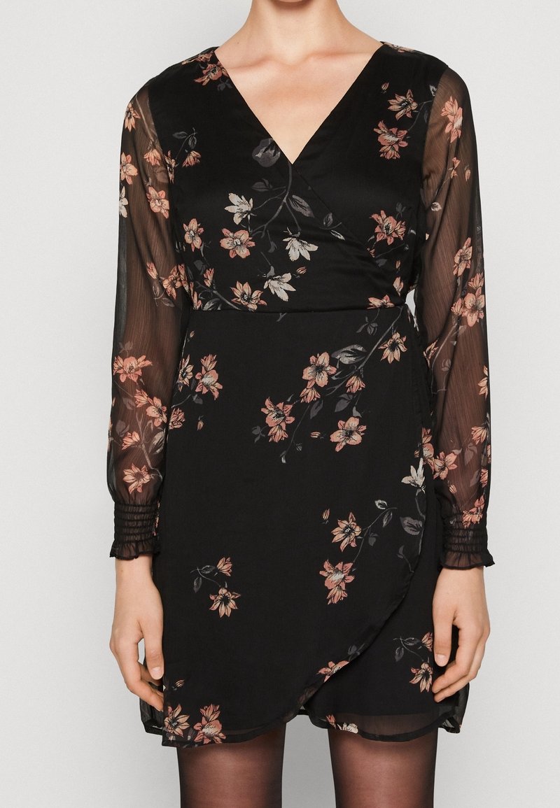 Robe portefeuille noire à col en V avec manches longues transparentes et imprimé floral rose, portée avec des collants noirs, montrée du cou à mi-cuisse.
