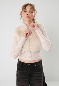 Vest cropped din blană sintetică bej, cu fermoar argintiu, purtat peste un top lung cu mânecă lungă de culoare roz deschis, asortat cu blugi negri cu talie înaltă.