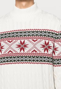 Bílý pletený rolák se červenými, černými a šedými geometrickými a hvězdicovými vzory na hrudi a horních pažích.