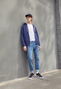 Marineblauer Reißverschluss-Hoodie über einem weißen T-Shirt, hellblaue Jeans und schwarze Sneakers mit weißen Sohlen, stehend gegen eine graue Wand.