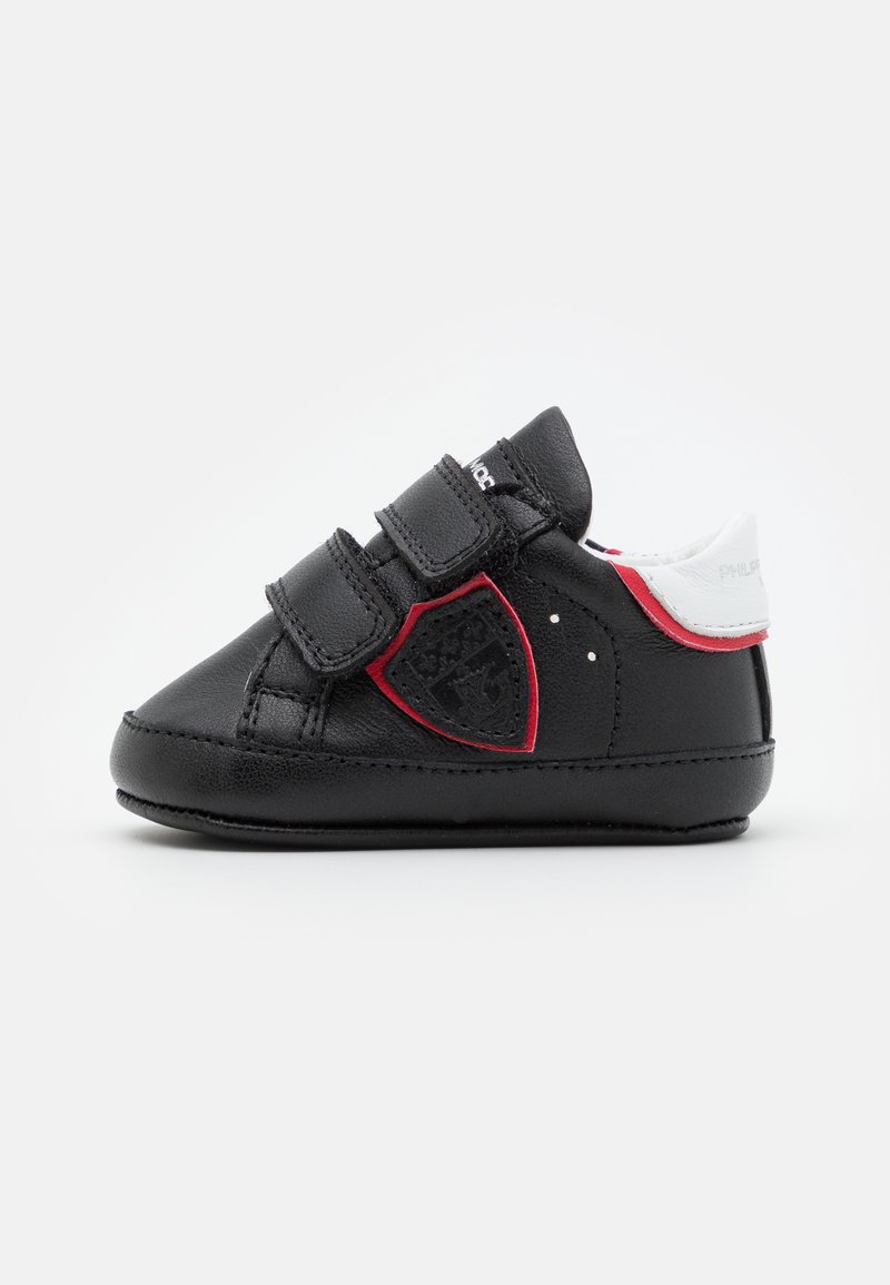 Sapatos de couro pretos para crianças pequenas com tiras de Velcro, detalhes em vermelho e um painel lateral texturizado com um design de escudo. Secção do calcanhar branca.